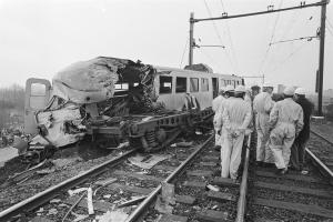Eine Schwarz-Weiß-Szene einer Zugkollision mit Menschen in Helmen, die herumstehen, Strommasten, Bäumen, Gebäuden und einem klaren Himmel im Hintergrund.