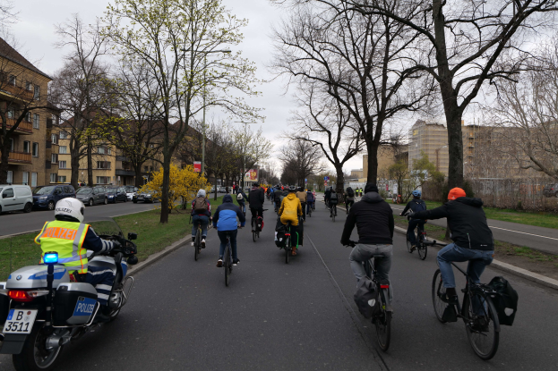 Eine Gruppe von Menschen, die auf Fahrrädern eine von Bäumen gesäumte Straße entlangfahren, begleitet von einem Polizisten auf einem Motorrad.