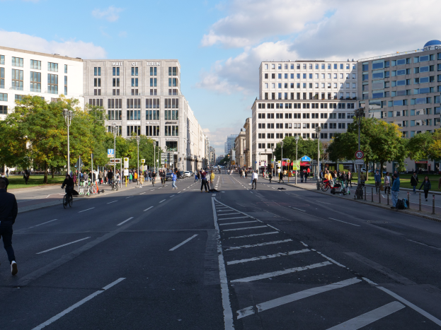 Eine belebte Straße in Berlin, Deutschland, mit Förderern und Radfahrern, hohen Gebäuden, Bäumen, Laternen und Schildern unter einem bewölktem Himmel.