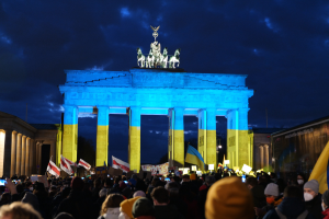 Eine Menschenmenge steht vor dem Reichstagsgebäude in Berlin, Deutschland, mit Fahnen und Plakaten in den Händen, mit einer Banner auf der rechten Seite und das Tor mit Statuen und Pfeilern geschmückt unter einem bewölktem Himmel.