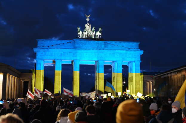 Eine Menschenmenge steht vor dem Reichstagsgebäude in Berlin, Deutschland, mit Fahnen und Plakaten in den Händen, mit einer Banner auf der rechten Seite und das Tor mit Statuen und Pfeilern geschmückt unter einem bewölktem Himmel.