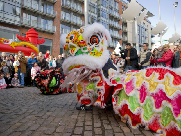 Ein lebendiges chinesisches Neujahrsfest in Amsterdam mit einem Löwen tanzen im Vordergrund und einer Menschenmenge drumherum, einige halten Kameras, vor einem Hintergrund aus Gebäuden, Laternenmasten und einem klaren blauen Himmel.