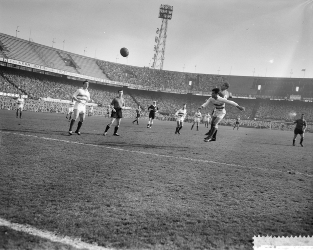 Eine Gruppe von Männern, die auf einem Feld mit Stadion, Flutlicht und einem klaren Himmel im Hintergrund Fußball spielen.