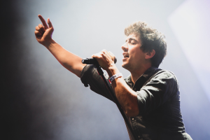 Ein Mann in einem schwarzen Hemd und Jeans singt in ein Mikrofon auf einer Bühne, mit geschlossenen Augen und einem jubelnden Publikum im Hintergrund.