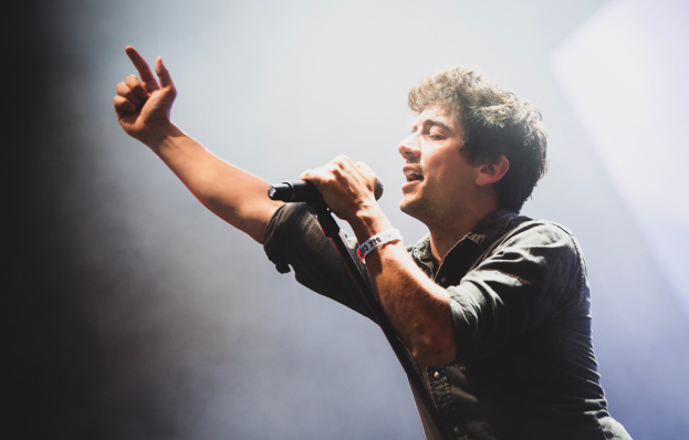 Ein Mann in einem schwarzen Hemd und Jeans singt in ein Mikrofon auf einer Bühne, mit geschlossenen Augen und einem jubelnden Publikum im Hintergrund.