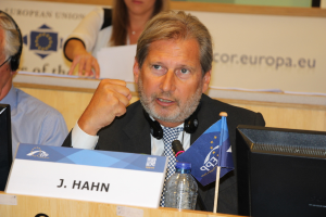 Ein Mann in einem schwarzen Blazer, weißem Hemd und schwarzer und weißer Krawatte spricht in ein Mikrofon mit einem Headset um den Hals, hinter einem Tisch mit einer Wasserflasche, Flagge, Laptop und Namensschild stehend; links von ihm arbeitet ein Mann in einem blauen Hemd an einem System und trägt Kopfhörer; dahinter hält eine Frau Papiere.