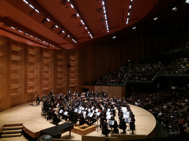 Ein großes Auditorium voller Menschen, die ein Konzert in der Royal Albert Hall in London besuchen, einige sitzen und halten Musikinstrumente, einige stehen auf der Bühne, Lichter an der Decke und eine Wand auf der linken Seite.