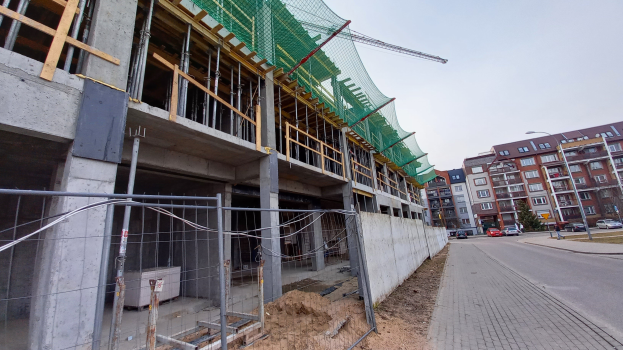 Großer Bau auf einer Straßenbaustelle, umgeben von Gebäuden, Straßenmasten, Straßenlaternen und Kraftfahrzeugen, mit einem klaren blauen Himmel.