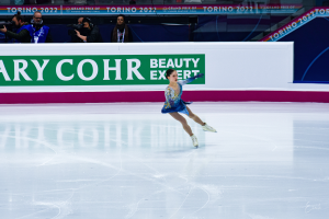 Eine Frau in einem blauen Kleid schlittert auf einer Eisbahn während eines professionellen Eiskunstlauf-Events, mit einer Menge von Menschen, die Kameras halten und ein Schild mit der Aufschrift "Tessa Virtue und Scott Moir" im Hintergrund.