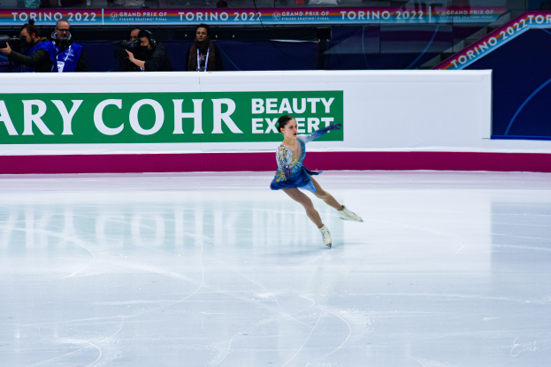 Eine Frau in einem blauen Kleid schlittert auf einer Eisbahn während eines professionellen Eiskunstlauf-Events, mit einer Menge von Menschen, die Kameras halten und ein Schild mit der Aufschrift "Tessa Virtue und Scott Moir" im Hintergrund.