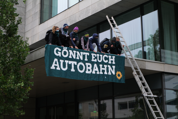 Eine Gruppe von Menschen steht auf dem Dach eines Gebäudes, hält eine Fahne, mit einem Baum links und Glasfenstern im Hintergrund, wahrscheinlich protestierend für die deutsche Regierung.