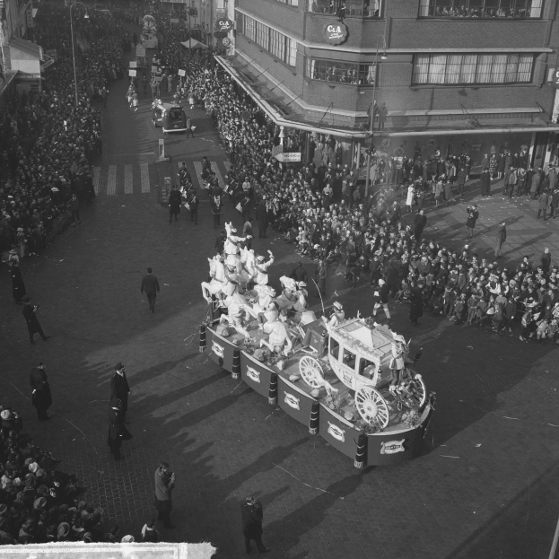 Eine große Menschenmenge umringt einen Festwagen bei einem Umzug, der mit bunten Luftballons und Bändern geschmückt ist, mit Gebäuden, Laternenmasten und Texttafeln im Hintergrund, in Schwarz-Weiß.