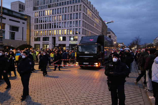 Eine Gruppe von Menschen steht vor einem Lastwagen auf einer Straße, umgeben von Gebäuden, Laternenmösten, Bäumen und einem bewölktem Himmel, einige tragen Mötzen und Masken, und ein Band mit einem Pfahl im Vordergrund.