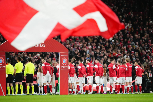Eine Gruppe von Menschen auf einem Fußballfeld mit einer roten und weißen Flagge im Vordergrund, einem Bogen mit Text "Bayern München vs Bayern Munich Betting Tips & Preview" im Hintergrund und einer großen Menge im Stadion.