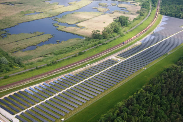 Luftaufnahme eines Solarparks mit Solarpanelen in einer grünen Wiese, umgeben von Bäumen und Wasser, mit einem Zug auf einer näheren Eisenbahnschiene.