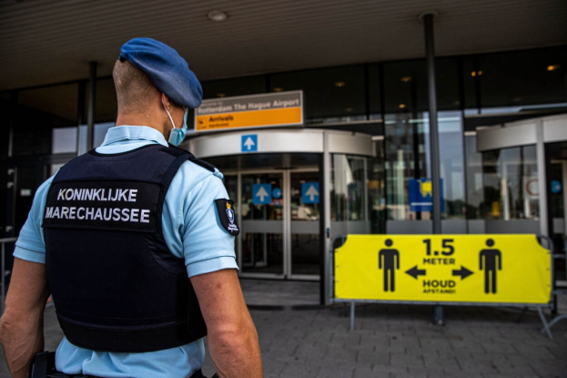 Ein Polizeibeamter in blauer Uniform und Maske steht vor einem Glasgebäude, mit gelben und anderen Informationsschildern im Hintergrund.