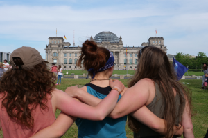 Drei Frauen in verschiedenen farbigen Kleidern, eine trägt eine Kappe, stehen vor dem Reichstag in Berlin, Deutschland, mit Menschen, Fahnen, Bäumen, Wolken und Himmel im Hintergrund.