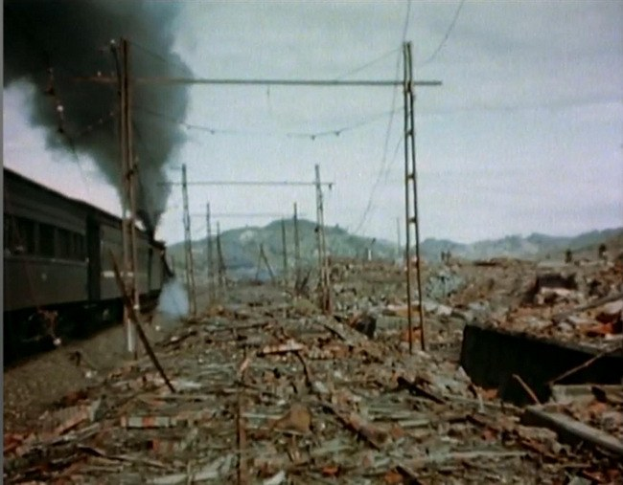 Ein Zug fährt durch ein beschädiges Gebiet mit Rauch, der aus ihm aufsteigt, umgeben von Masten und Drähten, Hügel und einem klaren Himmel im Hintergrund und verstreutem Schrott auf dem Boden.