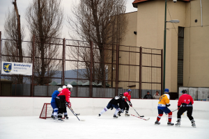 Menschen beim Eisschlittschuhlaufen auf einer Eisfläche mit Gebäuden, Bäumen, einer Straßenlaterne, einem Namensschild und Zäunen im Hintergrund unter einem klaren Himmel.