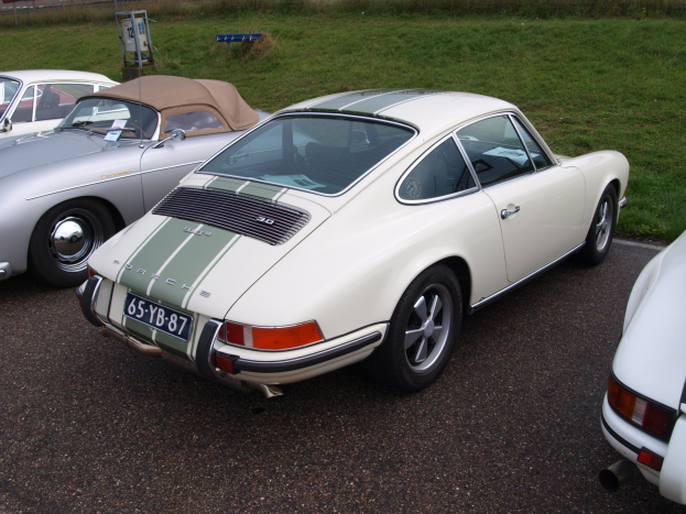 Ein weißer Porsche 911 auf einem Parkplatz mit anderen Autos, Gras, einem Schild, einem Zaun, Pflanzen, Bäumen und einem Gebäude im Hintergrund.