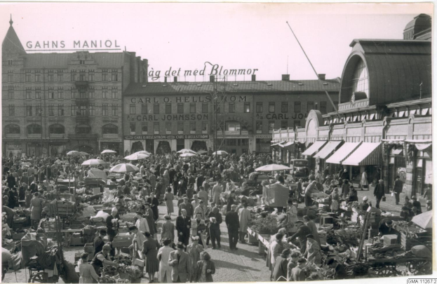 Schwarzes und weißes Foto eines belebten Berliner Markts mit Menschen, Gemüsekarren und Gebäuden im Hintergrund.
