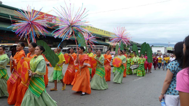 Eine Gruppe von Menschen in bunten Kostðmen geht eine Straße entlang, während eine Frau auf der rechten Seite eine Flasche hölt, und Fahrzeuge, Gebäude, Bäume, Mäste, Drähte und der Himmel im Hintergrund zu sehen sind.