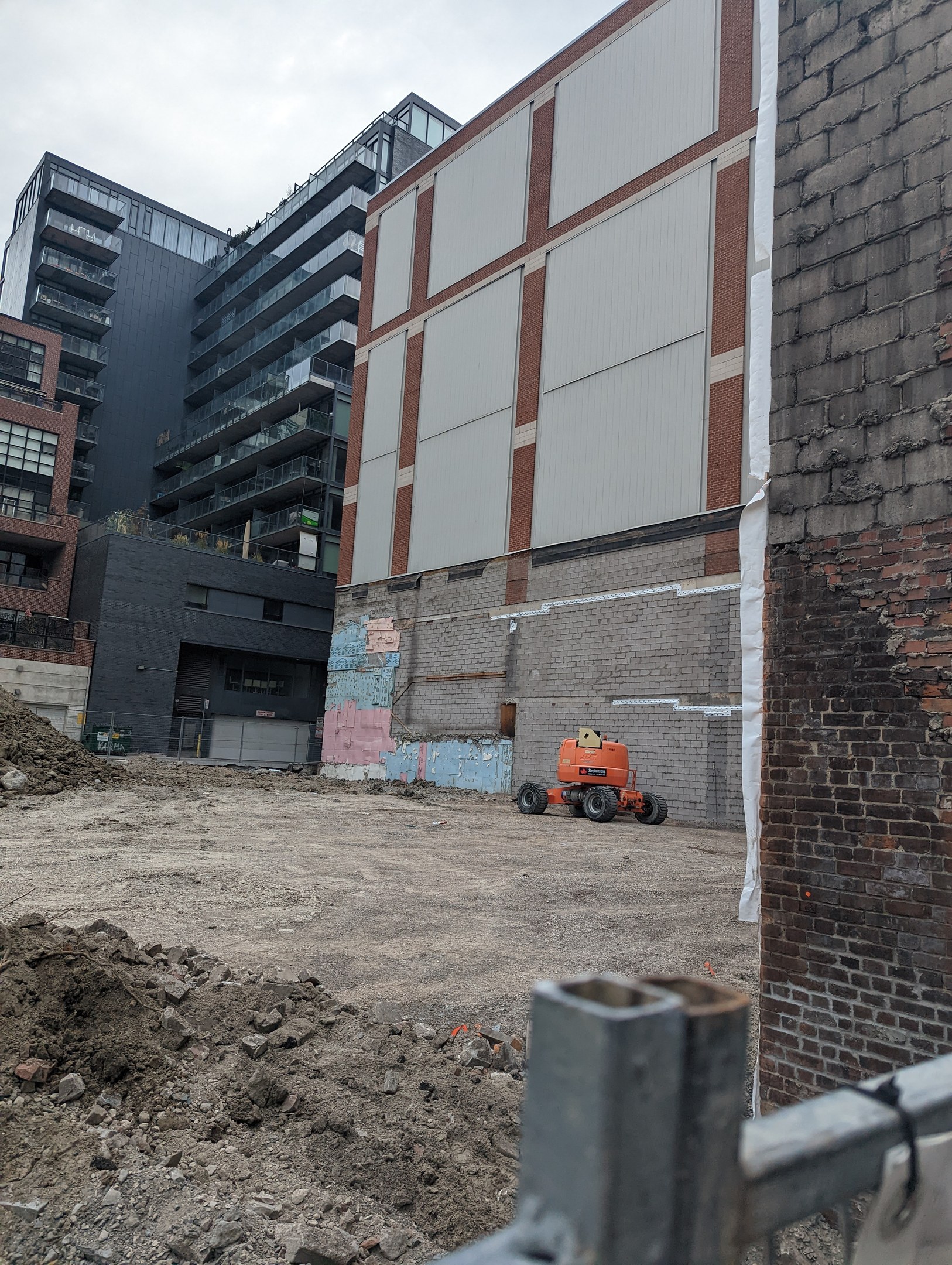 Baustelle mit einem großen Gebäude unter Abriss, einem Fahrzeug, einem Metallzaun und einem bewölkten Himmel.