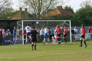 Fußballspieler sind in ein Fußballspiel auf einem Feld mit einem Tornetz involviert, während Zuschauer dahinter stehen, mit Bäumen und Häusern im Hintergrund.
