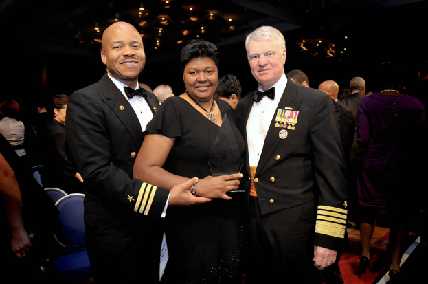 Zwei Männer in schwarzen Blazern und weißen Hemden stehen neben einer Frau in einem schwarzen Kleid, die eine Trophäe hält, auf einem roten Teppich, während andere sitzen und stehen und von beleuchteten Decken umgeben sind.