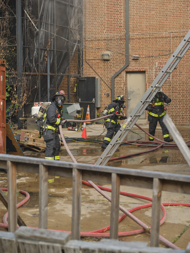 Eine Gruppe von Feuerwehrmännern in Helmen, die Rohre halten, arbeiten daran, ein Gebäude Feuer zu löschen, mit verschiedenen Ausrüstungen und Strukturen drum herum, einschließlich eines Metallzauns, Rohren auf dem Boden, einem Container, einem Verkehrskegel und einem Baum im Hintergrund.