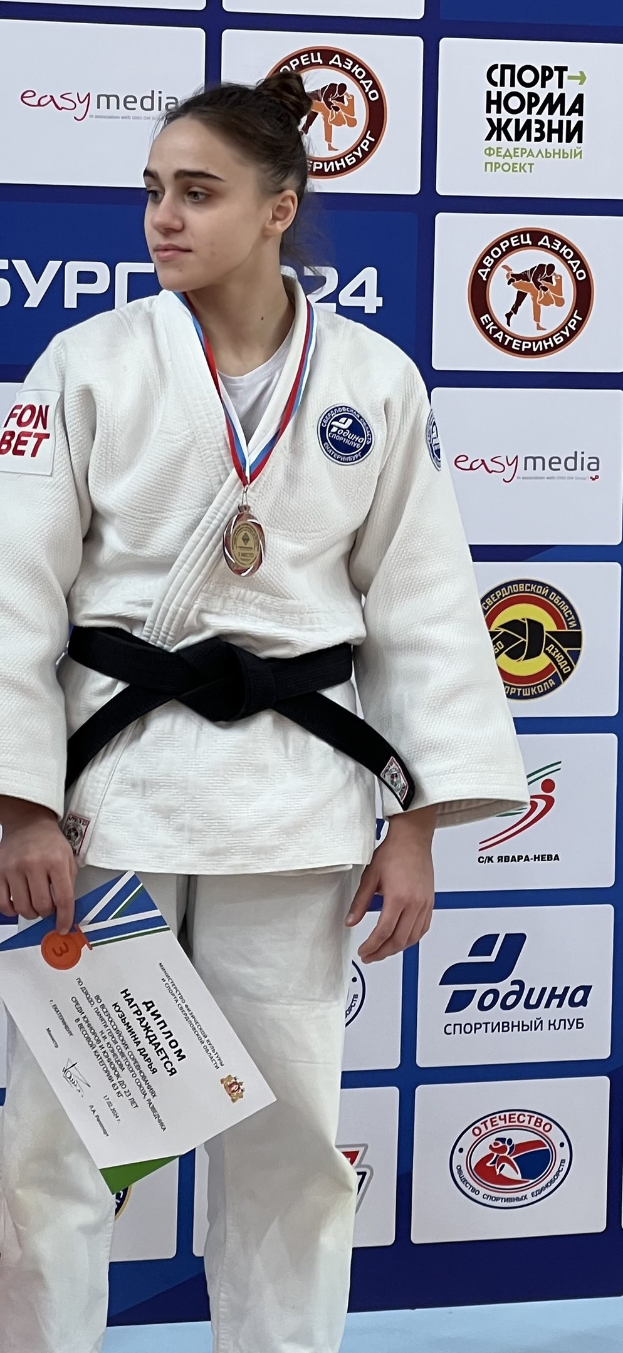 Frau in weißem Judo-Anzug mit schwarzem Gürtel steht auf einem Podium und hält eine Medaille und ein Zertifikat in den Händen, mit einem Werbetafel im Hintergrund.