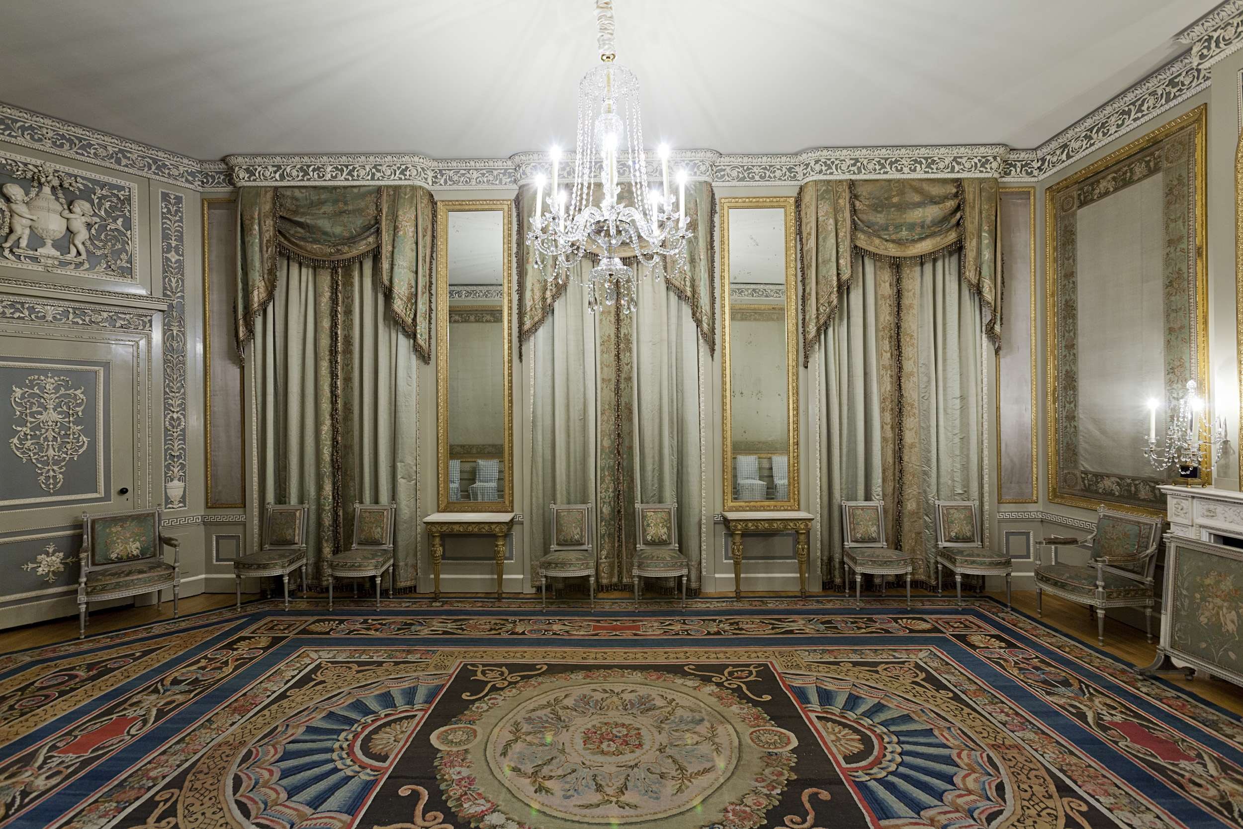 Ein luxuriöses Zimmer mit einem großen Teppich, Stühlen, Tischen, Spiegeln, Vorhängen, einer Deckenlampe und einem Kamin mit Kerzen auf der rechten Seite, mit kunstvoll verzierten Wänden.