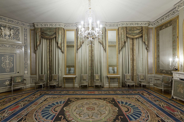 Ein luxuriöses Zimmer mit einem großen Teppich, Stühlen, Tischen, Spiegeln, Vorhängen, einer Deckenlampe und einem Kamin mit Kerzen auf der rechten Seite, mit kunstvoll verzierten Wänden.