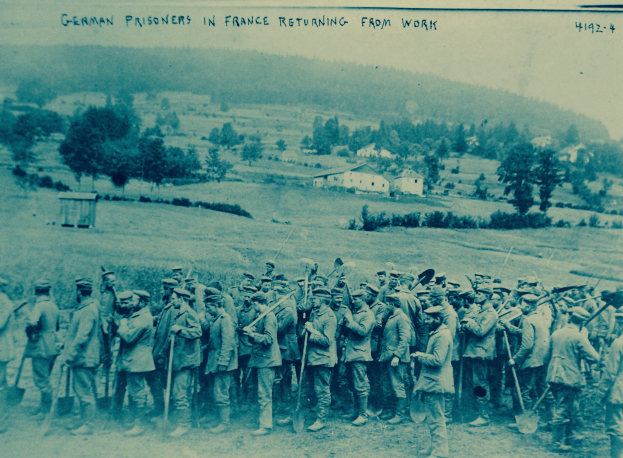 Eine Gruppe deutscher Gefangener in Frankreich mit Waffen im Bild, die auf dem Boden stehen, umgeben von Bäumen, Häusern und einem klaren blauen Himmel.
