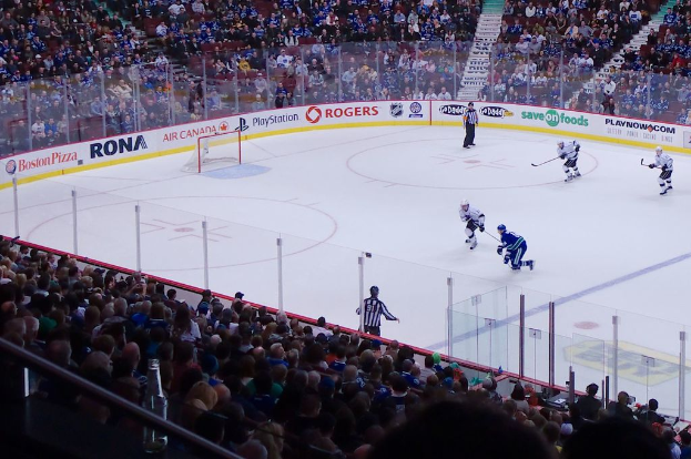 Vier Männer spielen Hockey auf einem Eisstadion, mit zwei in der Nähe des Tors und Zauns stehend, Zuschauern auf Stühlen im Vorder- und Hintergrund und einer Treppe im Hintergrund sichtbar.