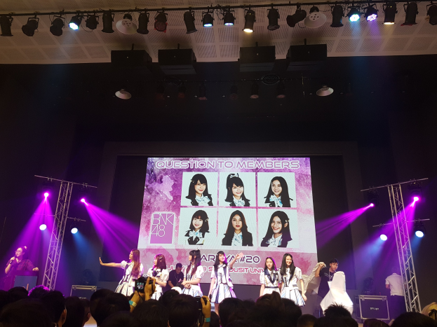 Eine Gruppe von Menschen tritt auf einer hell beleuchteten Bühne mit Trägern auf, wobei ein Sänger ein Mikrofon hält und ein Musiker ein Instrument spielt, während eine Menge unten das Ereignis aufzeichnet und ein großer Bildschirm Bilder der K-Pop-Gruppe BK48 anzeigt.