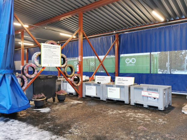 Ein gro├če Lagerhalle mit verschiedenen Reifen, Containern, Topfpflanzen, bedruckten Platinen, Polen, Lichtern, einem blauen Tuch und anderen Gegenst├Ąnden, mit B├Ąumen, Fahrzeugen und Schnee im Hintergrund.