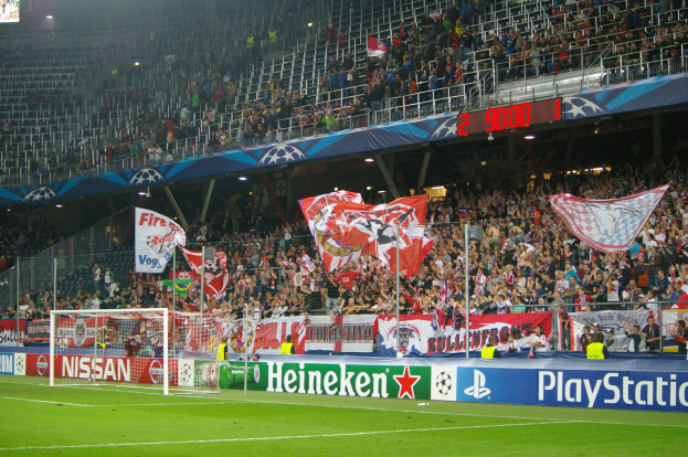 Ein Füßballspiel wird in einem hell erleuchteten Stadion mit einer großen Zuschauermenge, einem Tor in der Mitte, Bannern mit Text und Logos drumherum und einem Bildschirm in der oberen linken Ecke gespielt.