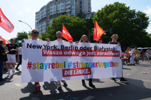 Eine Gruppe von Menschen geht eine von Bäumen gesäumte Straße in Berlin, Deutschland, entlang und hält ein Banner hoch, auf dem "Stay Rebel Stronger Together" steht, mit sichtbaren Gebäuden und Laternenpfählen und einem bewölkten Himmel.