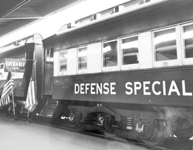 Schwarz-weißes Foto eines Zuges mit der Aufschrift "Defense Special" auf den Gleisen mit zwei Fahnen links und einer Decke oben.