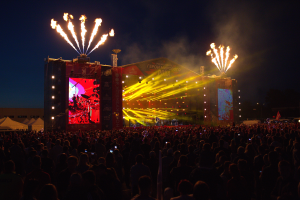 Eine große Menschenmenge versammelte sich auf einem Musikfestival mit einer Bühne in der Mitte, auf der Musiker im Scheinwerferlicht spielten und von Fahnen umgeben waren, Zelte links und ein dunkler Himmel mit Feuerwerk im Hintergrund.