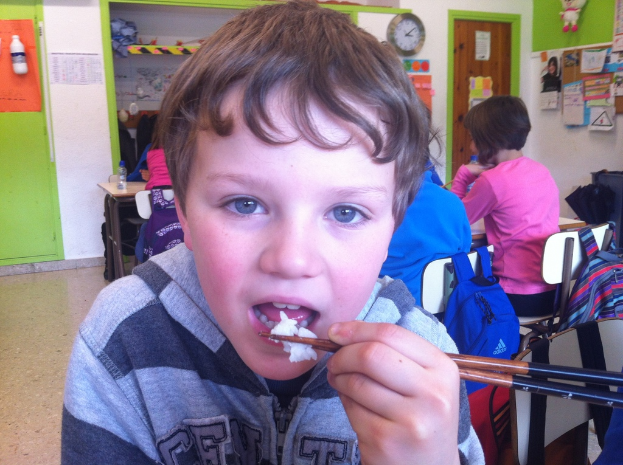 Ein Junge in einer grauen und schwarzen Jacke isst mit Stäbchen in einem Klassenzimmer, mit anderen Kindern hinter ihm, einer grün-weißen Wand, Lehrplänen und Postern an einer weißen Wand, einer braunen Tür und einer Wanduhr.