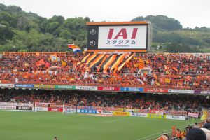 Ein Fußballspiel wird in einem Stadion mit einer großen Menge, saftigem Grün, einem Torpfosten in der Mitte, Bannern, Fahnen, einem großen Bildschirm, Bäumen und einem klaren blauen Himmel gespielt.