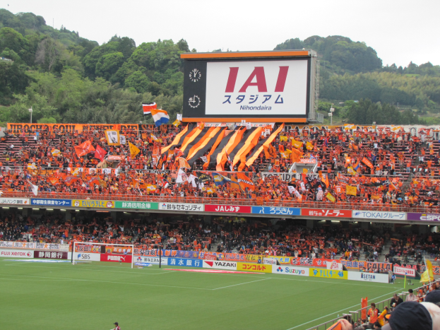 Ein Fußballspiel wird in einem Stadion mit einer großen Menge, saftigem Grün, einem Torpfosten in der Mitte, Bannern, Fahnen, einem großen Bildschirm, Bäumen und einem klaren blauen Himmel gespielt.