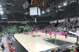 Ein Basketballspiel in einer großen Arena mit Zuschauern auf den Tribünen, Anzeigetafeln mit "Asvel vs Galatasaray" und einer beleuchteten Decke mit einem Bildschirm über dem Spielfeld.