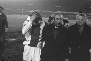 Eine Gruppe von Männern in Bedrängnis steht zusammen auf einem Fußballfeld, mit einem Stadion im Hintergrund, in einem Schwarz-Weiß-Bild.