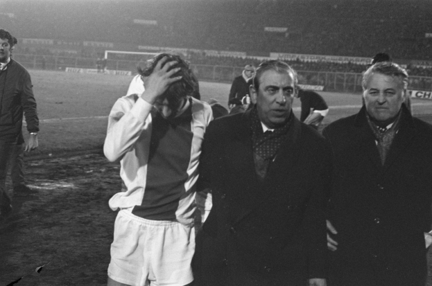 Eine Gruppe von Männern in Bedrängnis steht zusammen auf einem Fußballfeld, mit einem Stadion im Hintergrund, in einem Schwarz-Weiß-Bild.