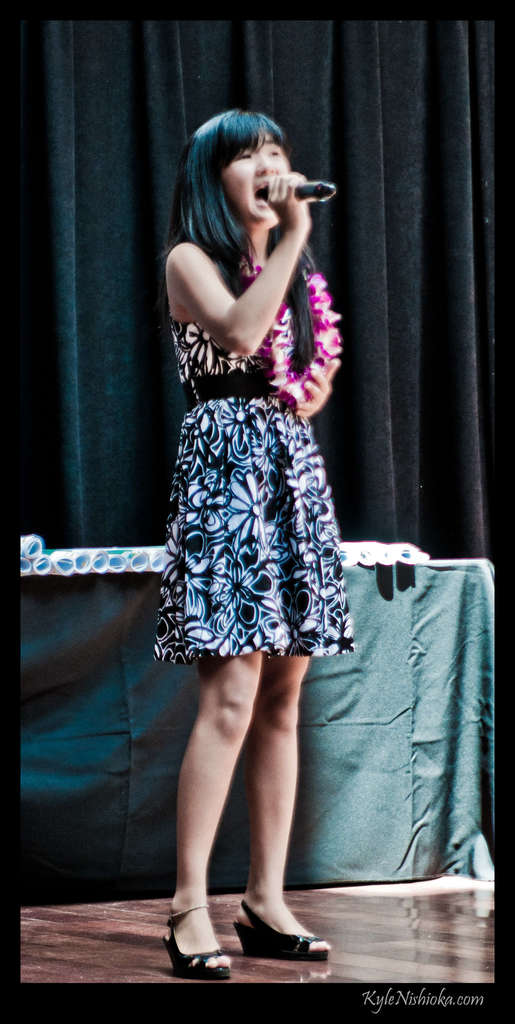 Eine Frau in einem bunten Kleid singt auf der B├╝hne in ein Mikrofon, mit einem Tisch mit Papieren und einem Vorhang im Hintergrund.