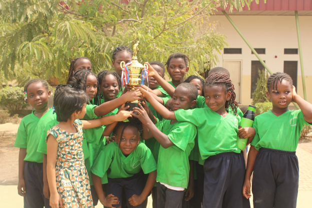 Gruppe von Kindern, die einen Pokal in einem Schulhof mit Bäumen und einem Gebäude im Hintergrund halten.