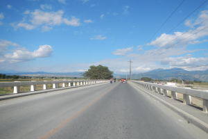 Eine Autobahn mit Motorradfahrern darauf, umgeben von Geländern, Strommästen mit Käblein, Bäumen, Hügeln und einem bewölkten Himmel.